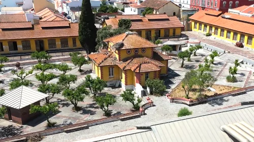 Vista aérea de uma cidade com edifícios com telhados laranja, um pátio com árvore e um grande castelo medieval em uma colina, cercado por vegetação e casas brancas, que mostra o impacto da reconstrução de Fábrica do Inglês na área.