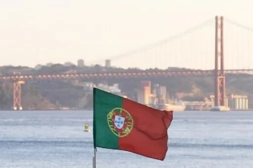 Durante la giornata nazionale del portogallo una bandiera portoghese sventola in primo piano con il ponte 25 aprile di Lisbona sullo sfondo sul fiume Tejo. Lo scenario include strutture sfocate sulla sponda opposta.