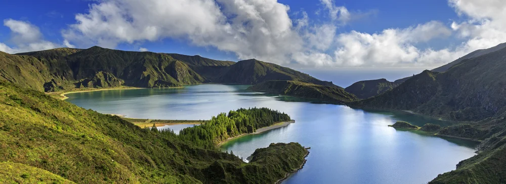 Vista aérea açoriana da Lagoa do Fogo, um lago azul imaculado rodeado por um terreno verde e montanhoso sob um céu parcialmente nublado. O lago possui várias penínsulas e ilhas cobertas por uma vegetação exuberante e o cenário geral é sereno e natural.